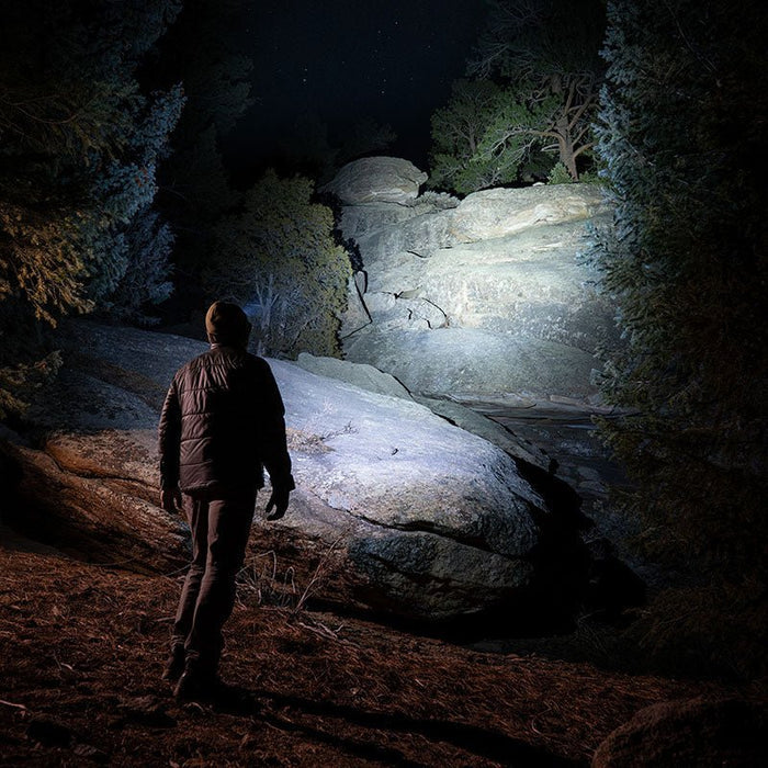 Person walking through a dark forest illuminated by a 1200 lumen Fenix HM55R RENEGADE headlamp on a rocky path.