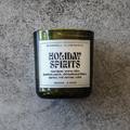 A Burnwell Illuminaries 'Holiday Spirits' scented candle in a dark green glass jar with a white label, placed on a textured gray stone surface.